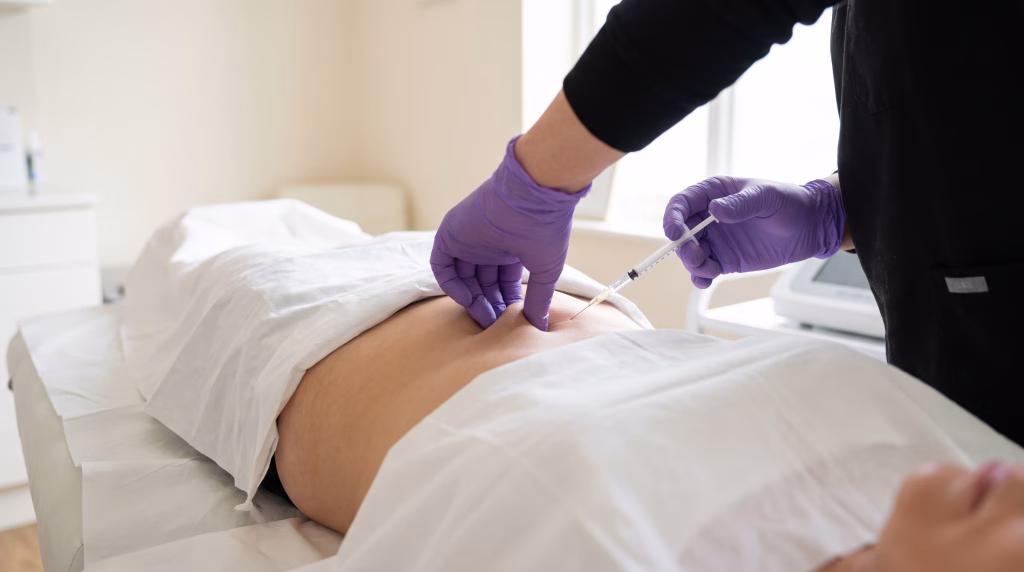 Gloved hands in black scrubs lifting a small pocket of lower-stomach skin and placing a fine-needle fat dissolving injection into it